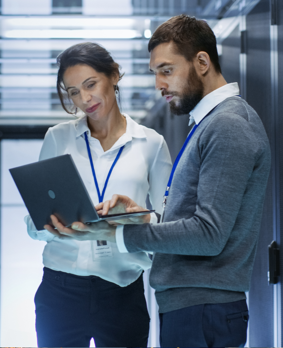 A man and woman in a server room. The man is showing the woman something on his laptop.