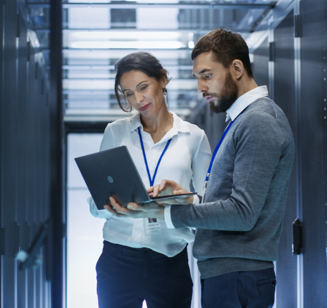 A man and woman in a server room. The man is showing the woman something on his laptop.